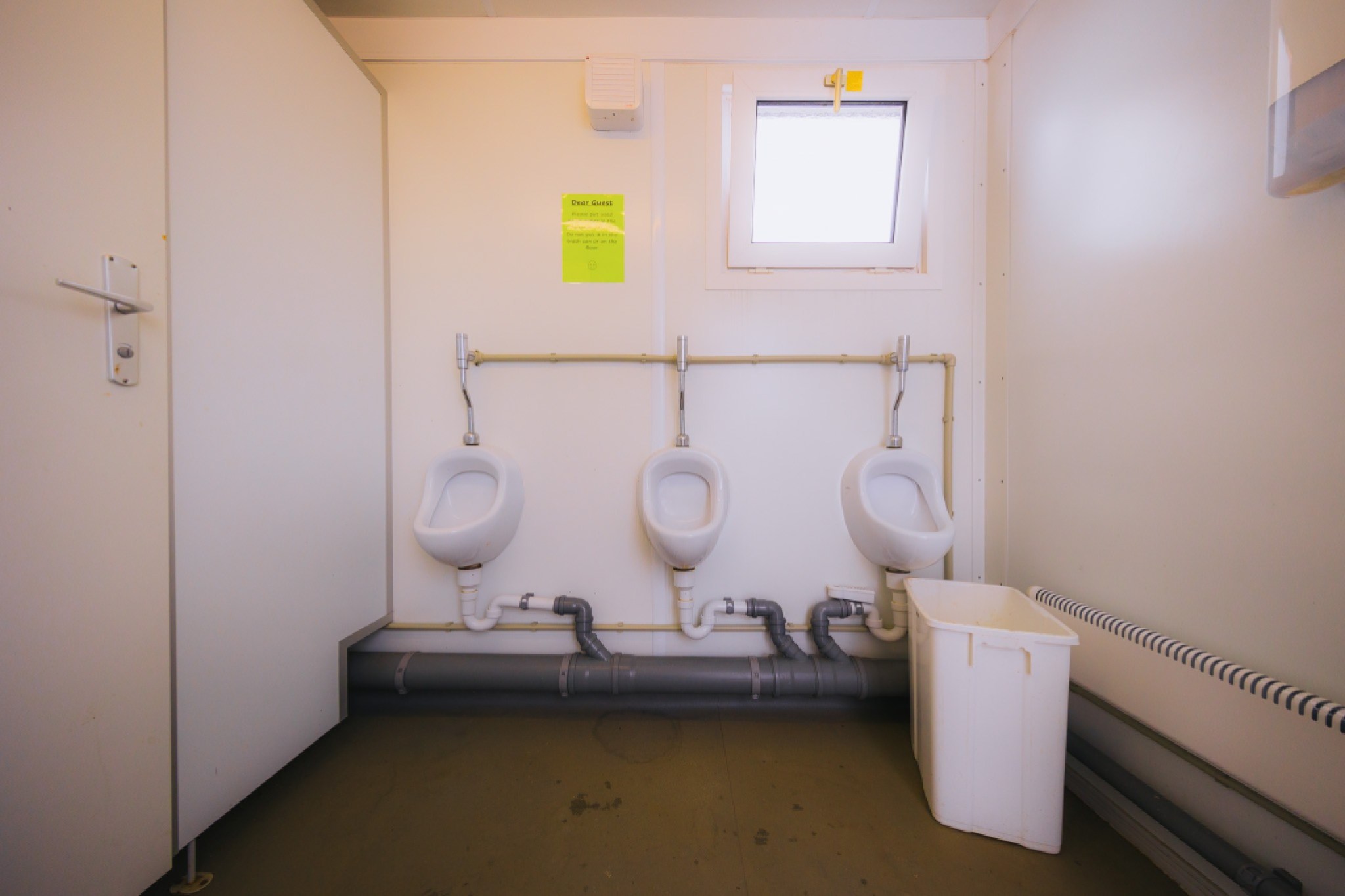 Three white urinals are mounted on a wall in a small restroom. Above them, a window and a green notice are visible. A white trash bin is on the right.
