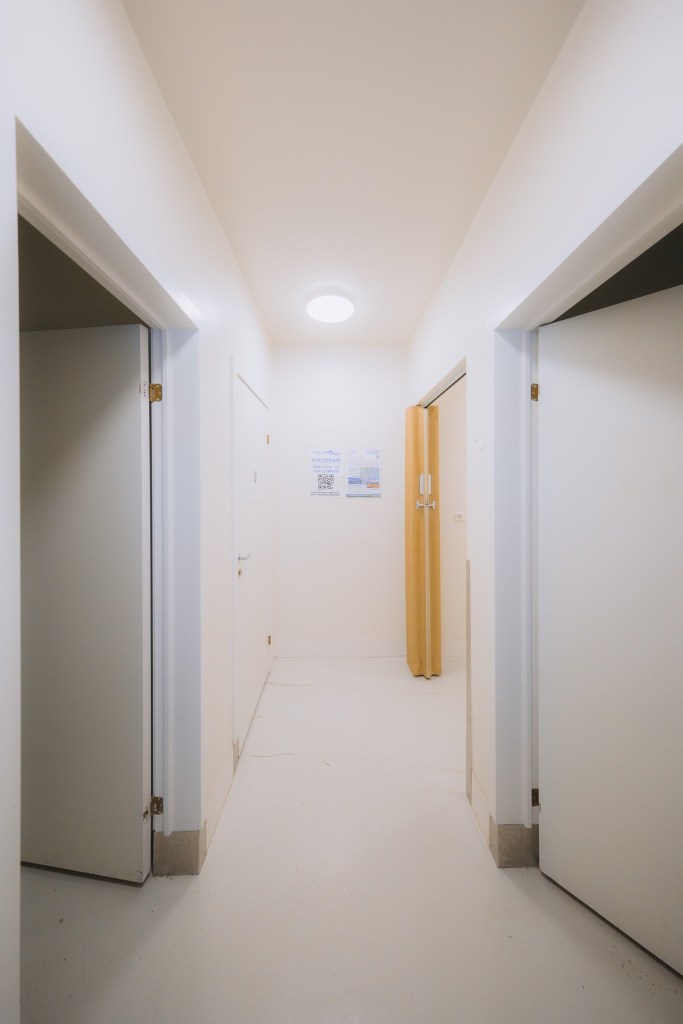 Minimalist hallway with three white doors, bright ceiling light, and beige partition curtain. Posters with text are on the wall.