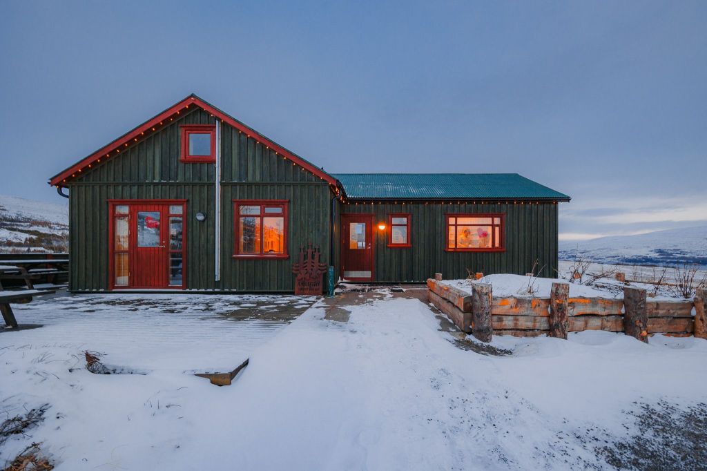 Cozy cabin with glowing windows in snowy landscape, dusk light. Rustic wooden structure, festive string lights, and serene winter atmosphere.