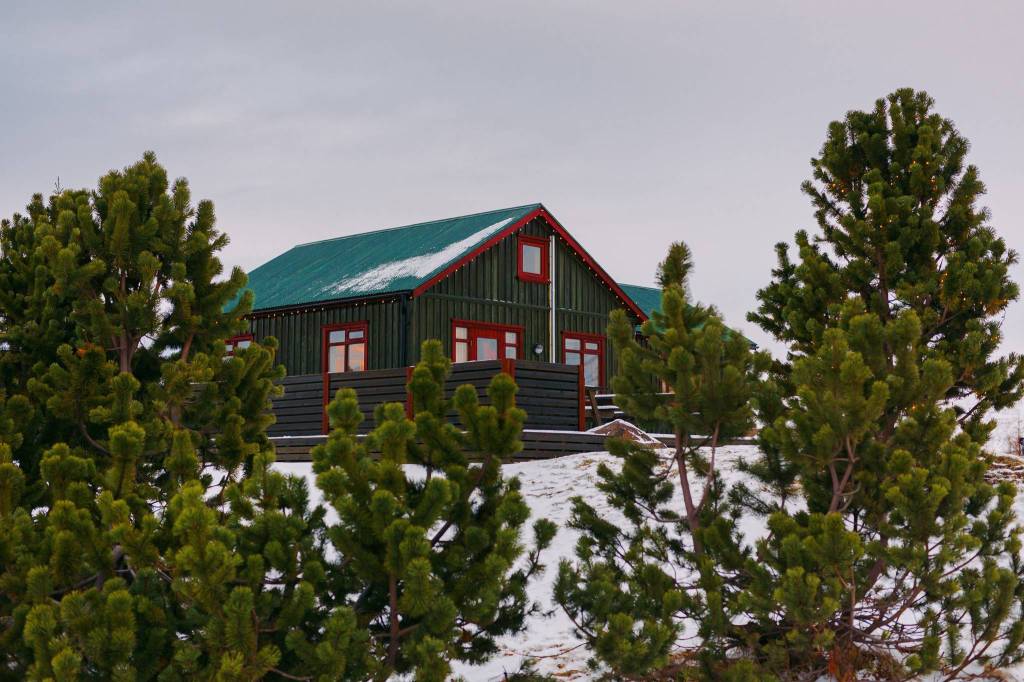 A cozy dark green cabin with a teal roof and red accents sits amid snowy ground, surrounded by evergreen trees, under a cloudy sky.