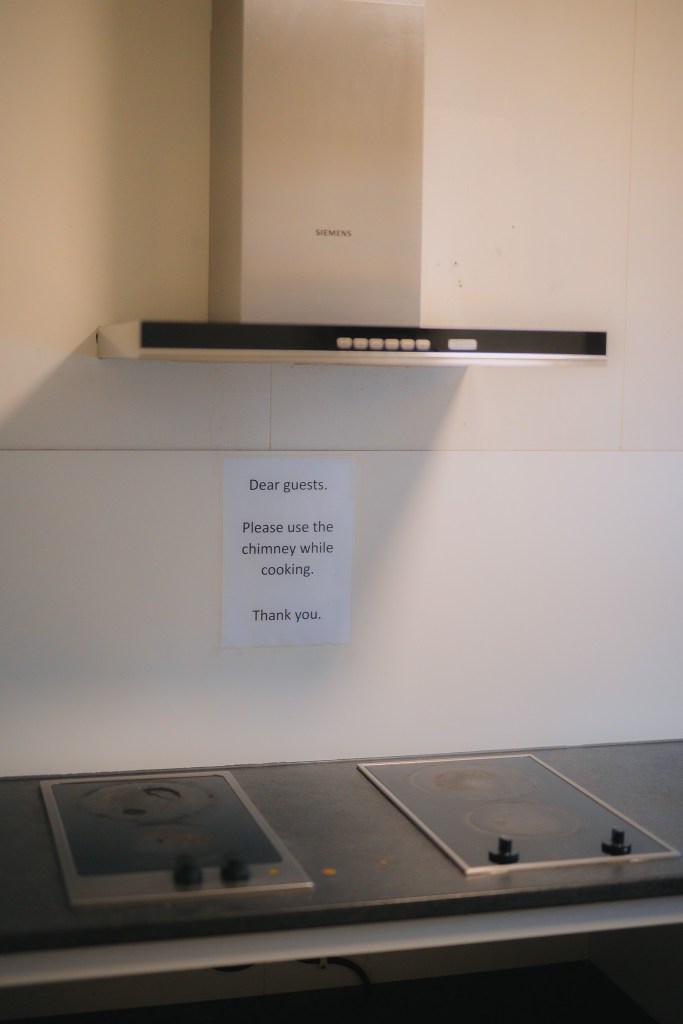 Kitchen scene with a modern range hood above an electric stovetop. A note instructs guests to use the chimney while cooking, conveying a polite request.