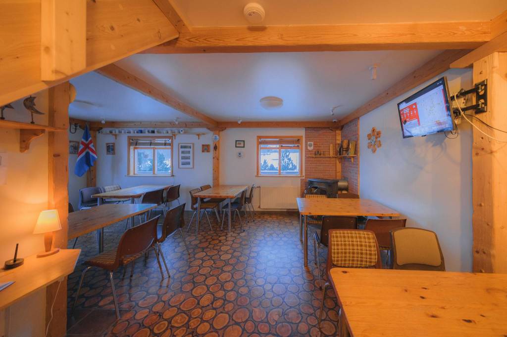 Cozy wooden dining area with a warm glow, featuring scattered tables and chairs. Wall adorned with a flat-screen TV and decorations.
