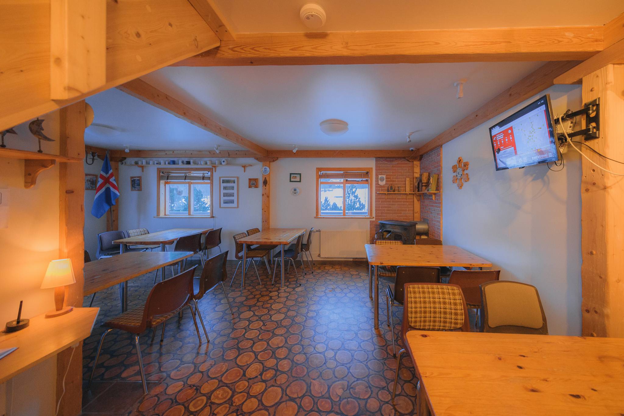 Cozy wooden dining area with a warm glow, featuring scattered tables and chairs. Wall adorned with a flat-screen TV and decorations.