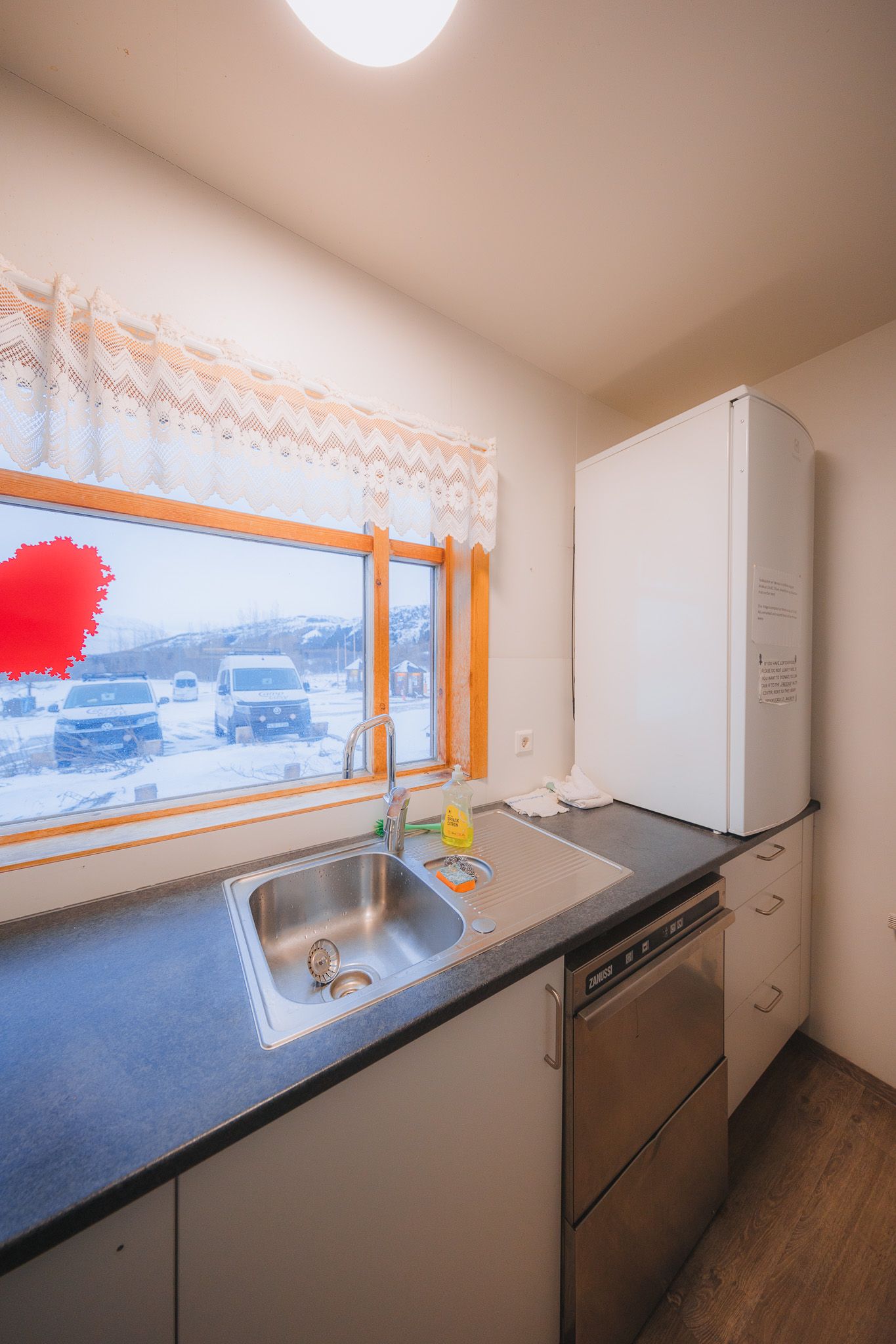 Compact kitchen with a sink, dishwasher, and fridge. A window with lace curtains shows a snowy scene outside.