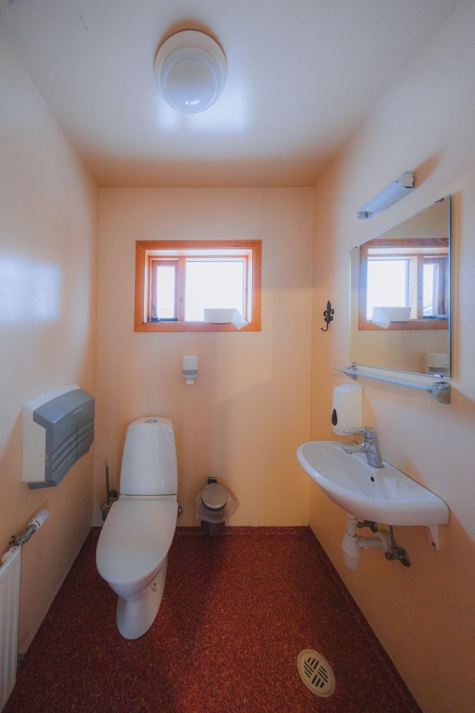 Small bathroom with cream walls and a red speckled floor. Includes a toilet, a wall-mounted sink, a rectangular mirror, and a small window.