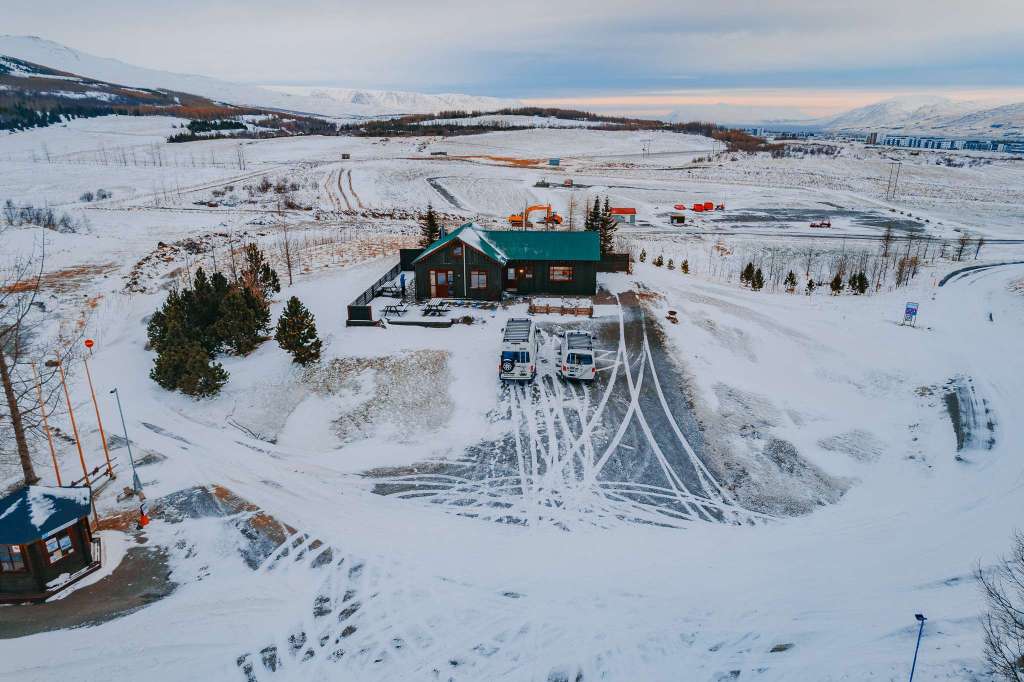 A snow-covered landscape with a dark green-roofed cabin at the center, surrounded by tire tracks. Two campervans are parked nearby.