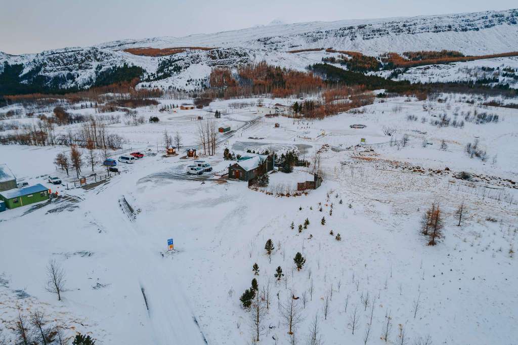 Aerial view of a snow-covered rural area with scattered cabins, parked vehicles, and sparse trees. Mountains are visible in the background under a cloudy sky.