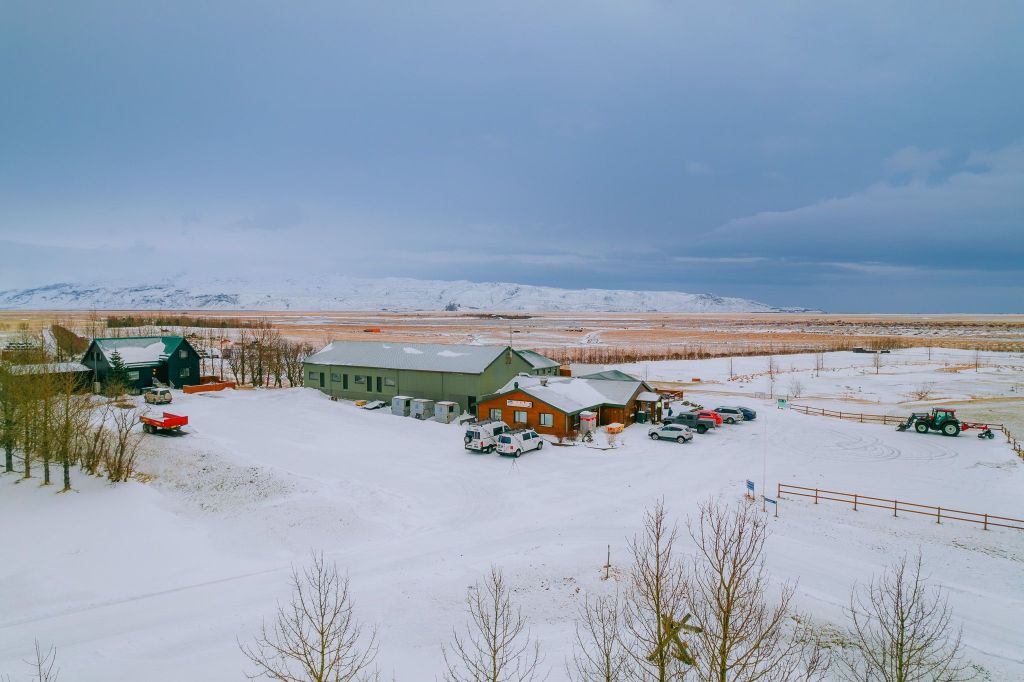 A snowy landscape features a cluster of buildings surrounded by leafless trees. Several vehicles are parked nearby, with snow-capped mountains in the distance.