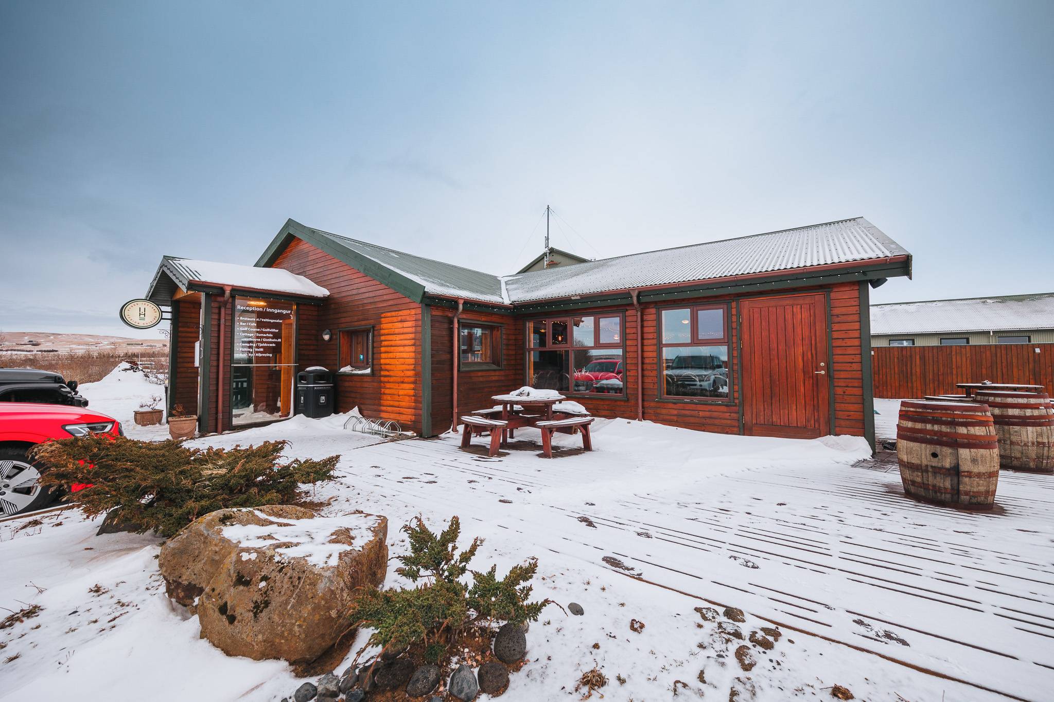 Log cabin in snowy landscape with a wooden picnic table and barrels, surrounded by light snow, under a cloudy sky.