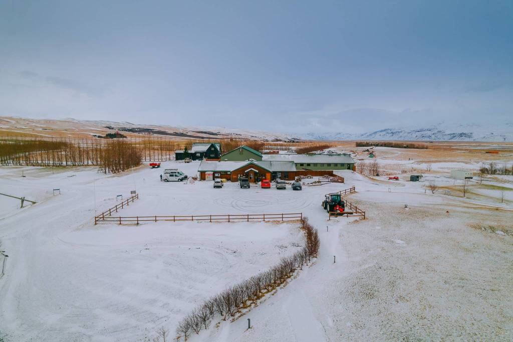 A snow-covered landscape featuring a large building surrounded by parked cars and trees. A tractor is visible, with distant hills under a cloudy sky.