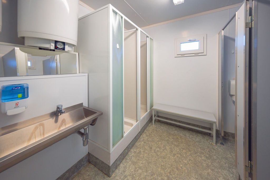 Clean, modern shower room with a metal sink, hand sanitizer, two shower stalls, a small window, and a bench.
