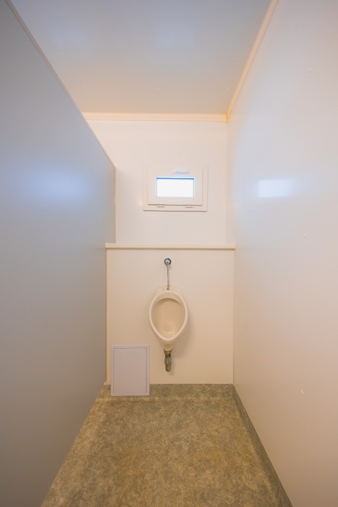 A small, minimalist restroom with white walls features a urinal mounted on the back wall beneath a small window.