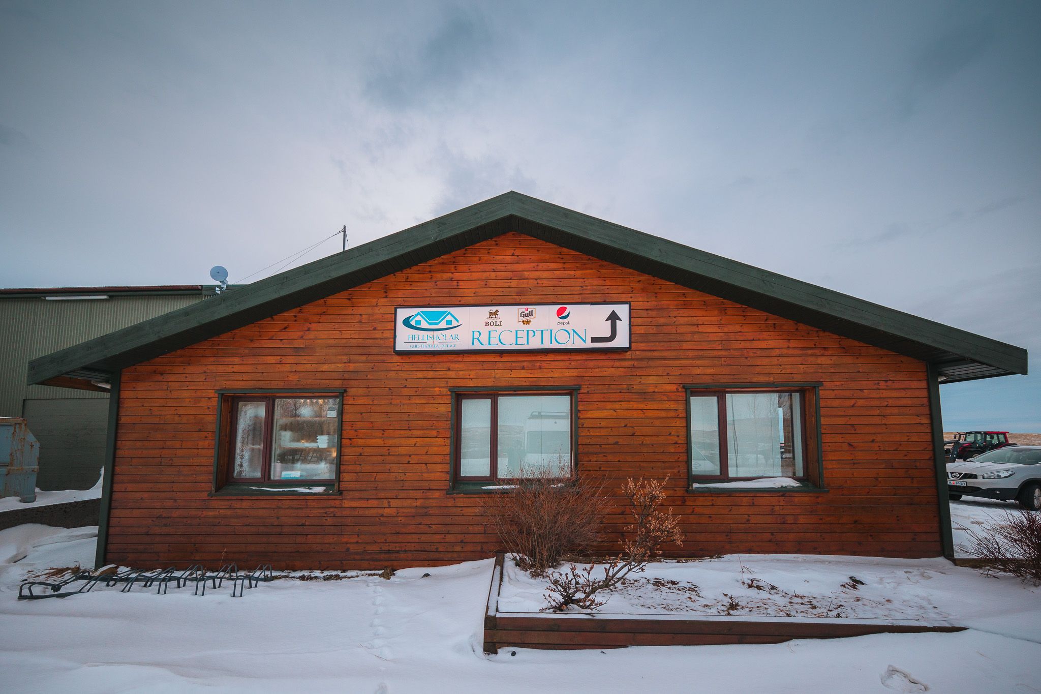 Wooden cabin with a “Reception” sign, surrounded by snow. Three windows on the front, creating a welcoming winter atmosphere against a cloudy sky.