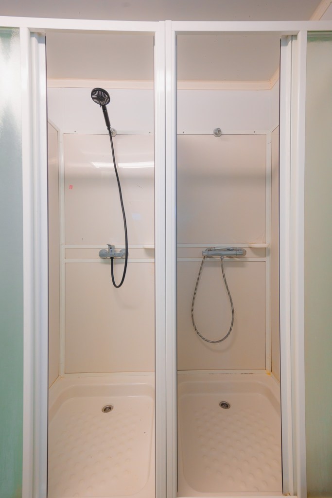 Two adjacent white shower stalls with sliding glass doors. Each has a handheld showerhead, one on the left with a black hose.