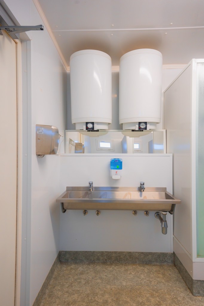 Stainless steel sink with two taps in an industrial bathroom. Two large water heaters above, a soap dispenser, and a hand dryer.