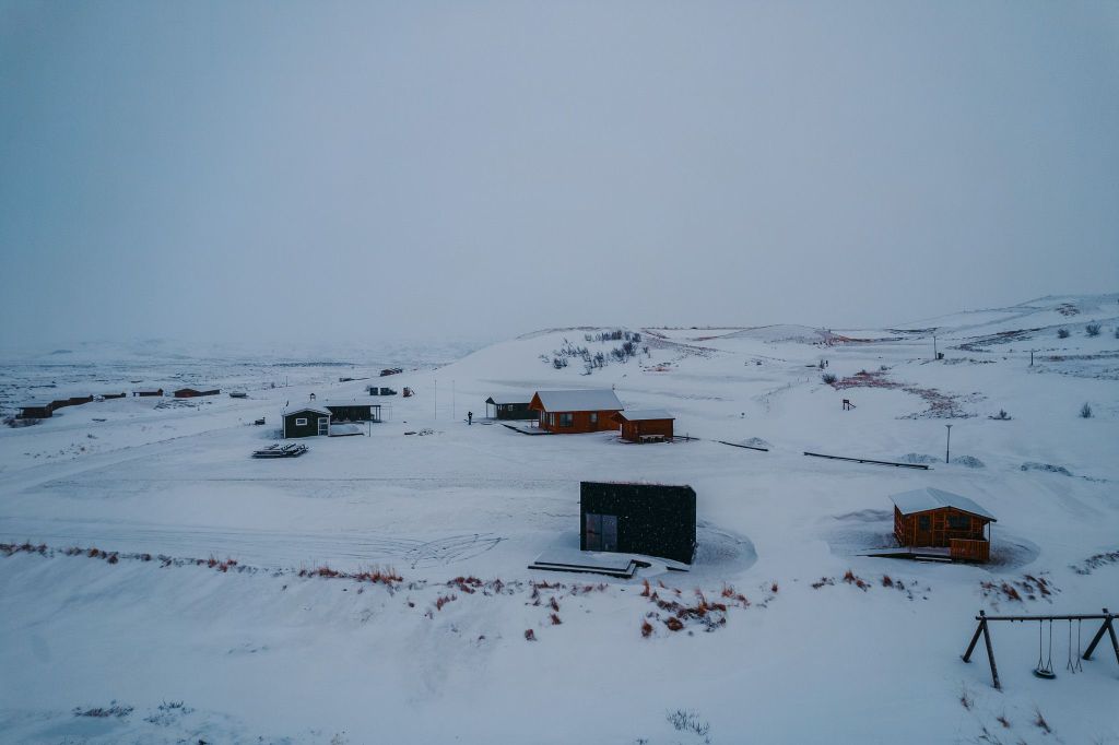 Snow-covered landscape with scattered wooden cabins and a swing set under an overcast sky.