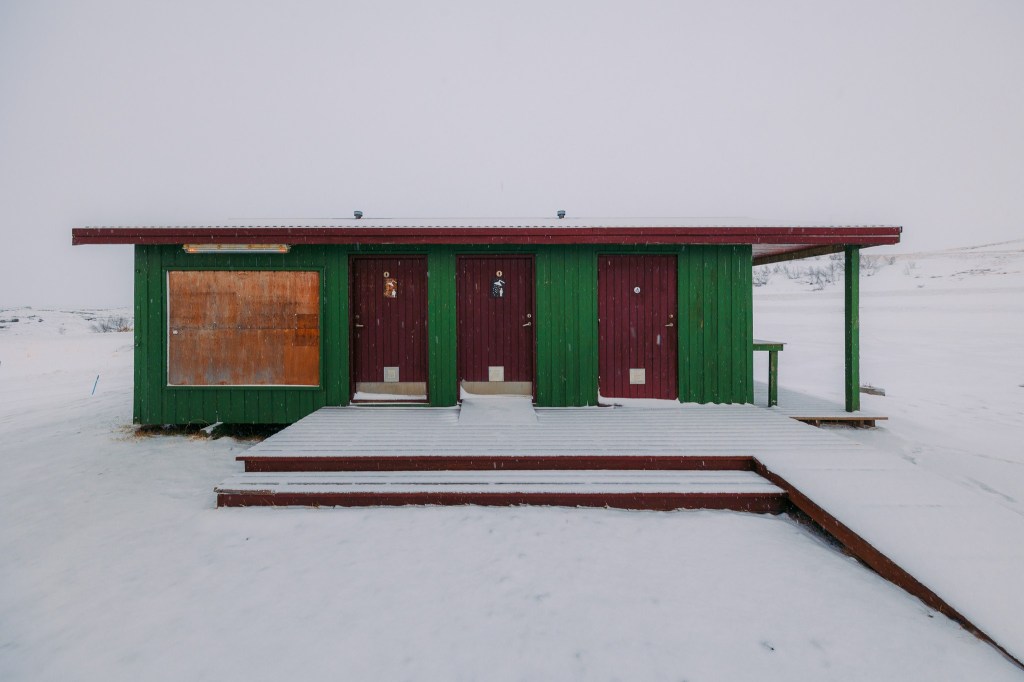 A small green and red cabin with three doors stands isolated in a snowy landscape.