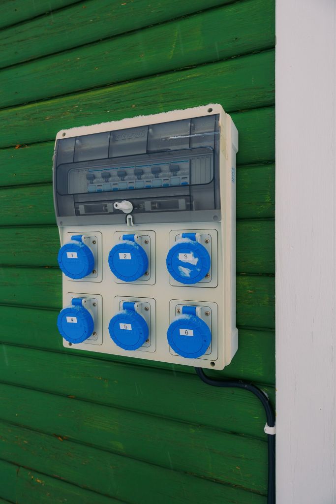 An electrical panel with six blue socket outlets is mounted on a green wooden wall. The panel has a transparent cover and numbered sockets.
