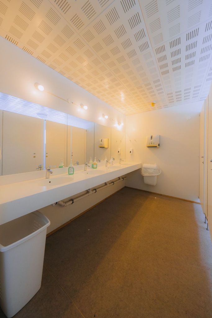 A clean, brightly lit public restroom with long, white countertops, multiple sinks, soap dispensers, mirrors, and a trash bin.