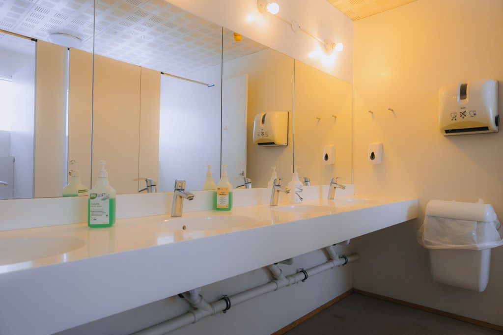Public restroom with a long white counter, four sinks, and mirrors. Bright lighting, soap dispensers, and paper towel holders.