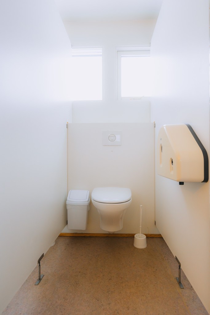 A clean, minimalistic bathroom with a white toilet, small trash bin, toilet brush, and wall-mounted paper dispenser.