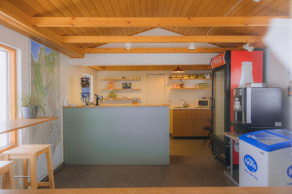 A cozy cafe interior with a wooden ceiling, bar counter, and stools. There’s a Coca-Cola fridge, ice cream freezer, and coffee machine. Shelves display snacks and decor.