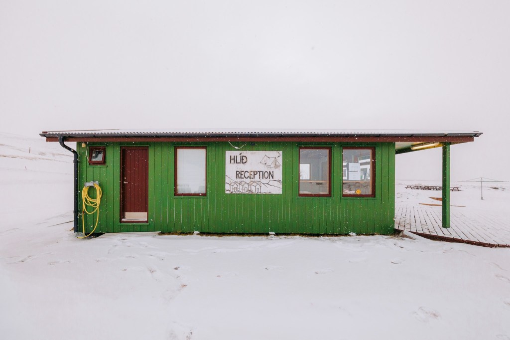 Green building with "Hlíð Reception" sign, surrounded by snow. The structure has a brown door, three windows, and a coiled yellow hose on the left.