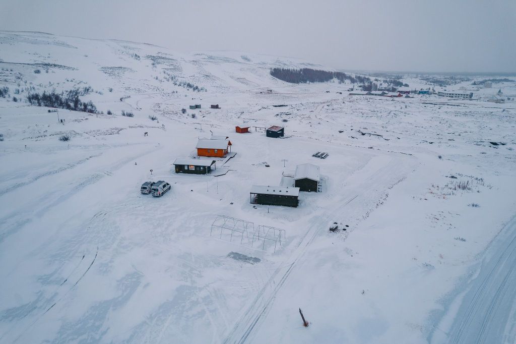 Aerial view of a snowy landscape with scattered buildings, vehicles, and distant hills.
