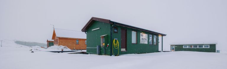 A green building on a snowy field with a wooden cabin and another structure in the background.