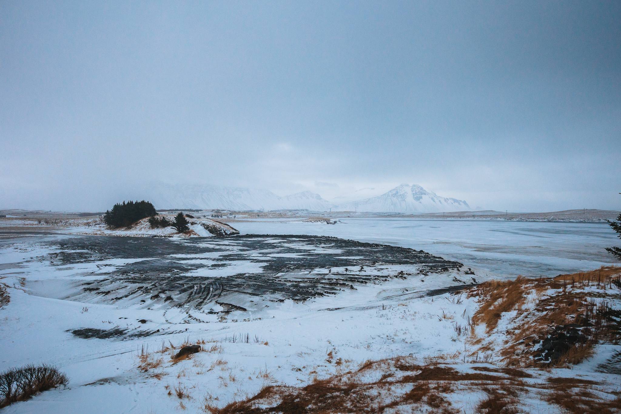 Snow-covered landscape with frosty grass and a frozen lake under a gray, overcast sky. Mountains are faintly visible in the distant background.