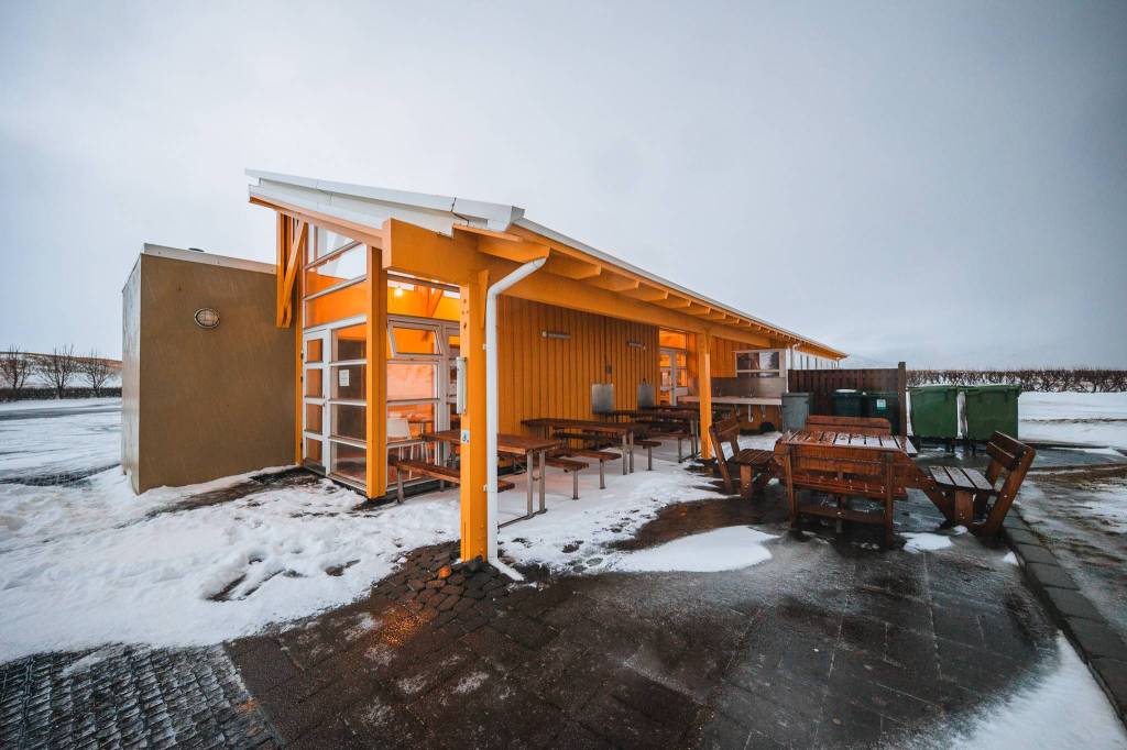 A cozy yellow cabin with large windows glows warmly in cold, snowy weather. Wooden benches and tables are outside, set against a cloudy, gray sky.