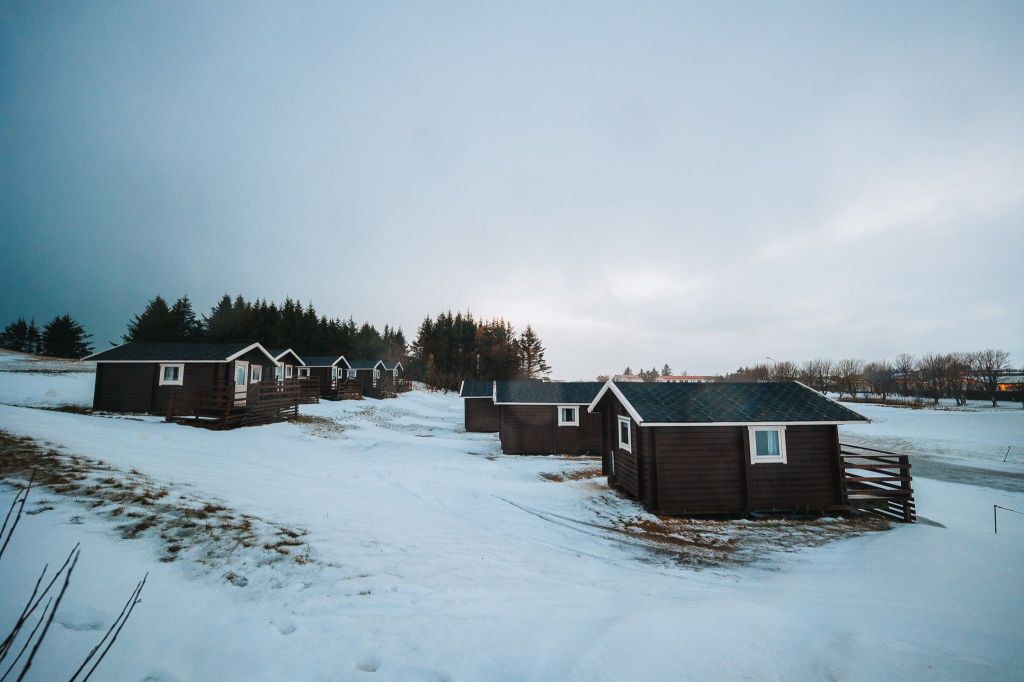 Row of small brown cabins on a snowy landscape, surrounded by sparse trees and overcast sky.