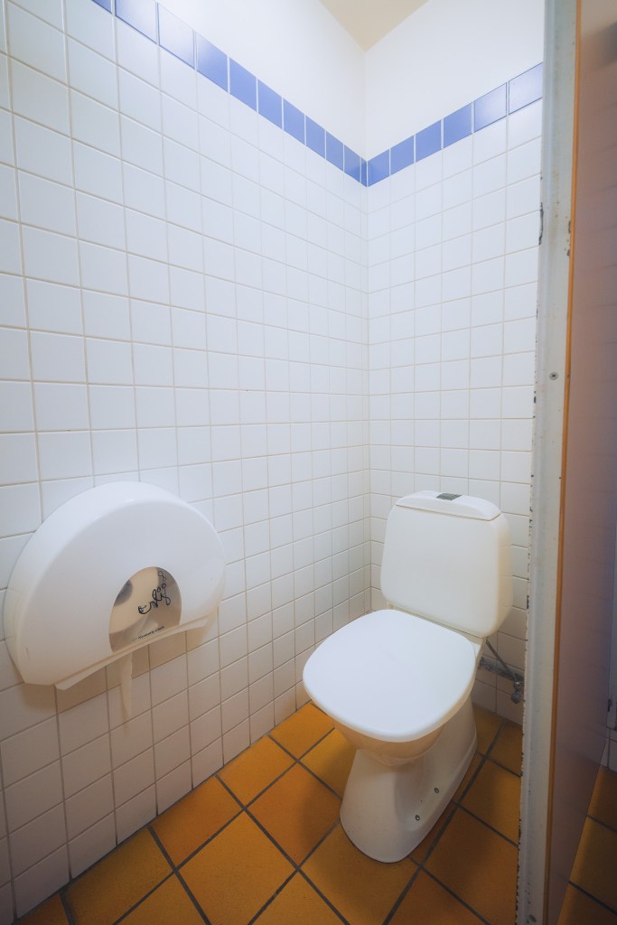 A clean public restroom with white tiled walls and blue trim. There is a white toilet and a large wall-mounted toilet paper dispenser, set on an orange-tiled floor.