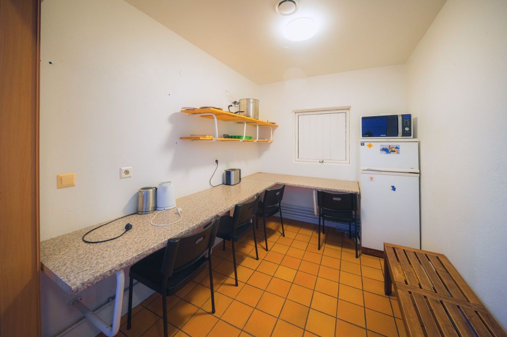 Small kitchen with a tiled floor, long granite countertop, and four black chairs. A white fridge and microwave are on the right, shelves above.