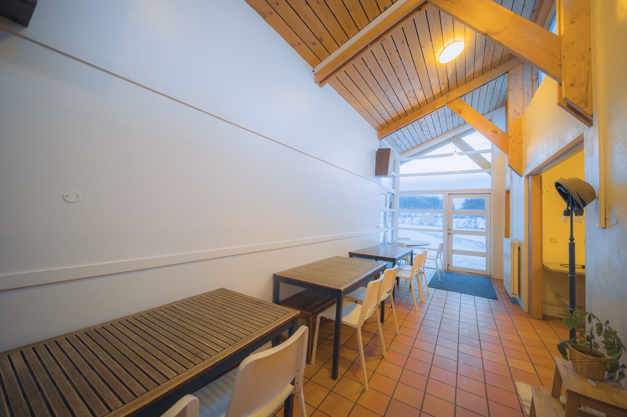 A cozy room with a slanted wooden ceiling and large windows overlooking a snowy landscape. Wooden tables and white chairs line the tiled floor.