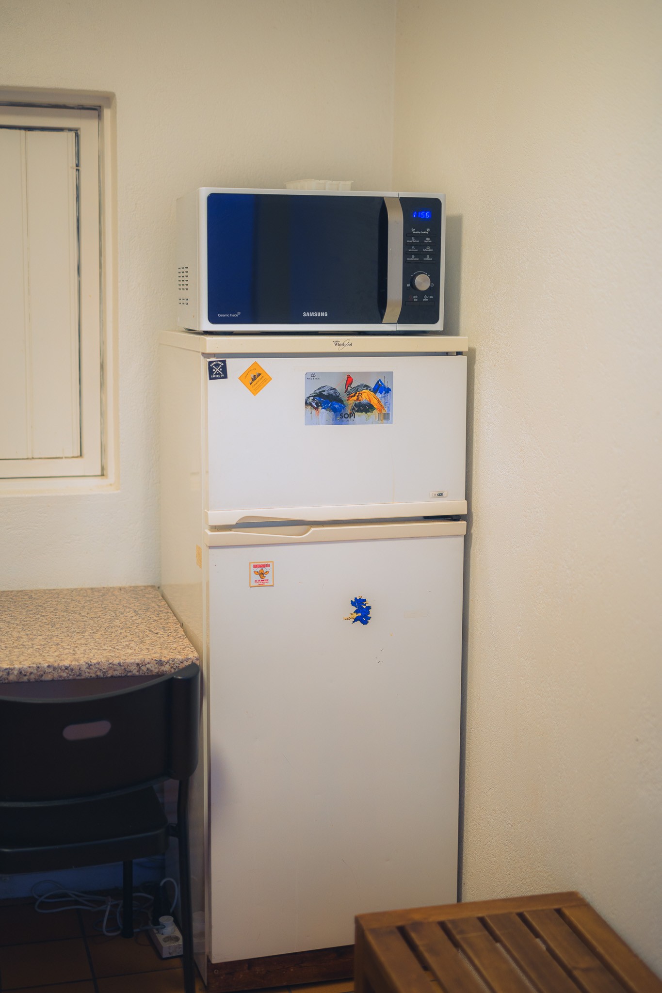 A white refrigerator with colorful magnets stands against a light wall. A black microwave sits atop it. Nearby, a small counter with a chair.