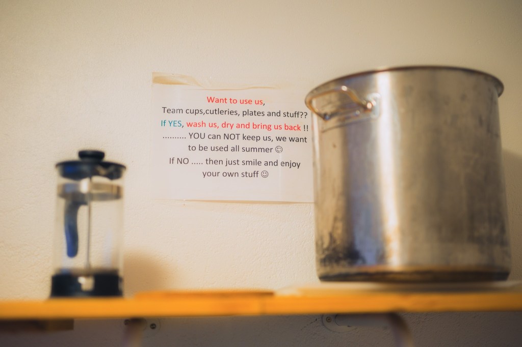 A large metal pot and a French press sit on a wooden shelf. A humorous sign on the wall instructs users to wash and return kitchen items.