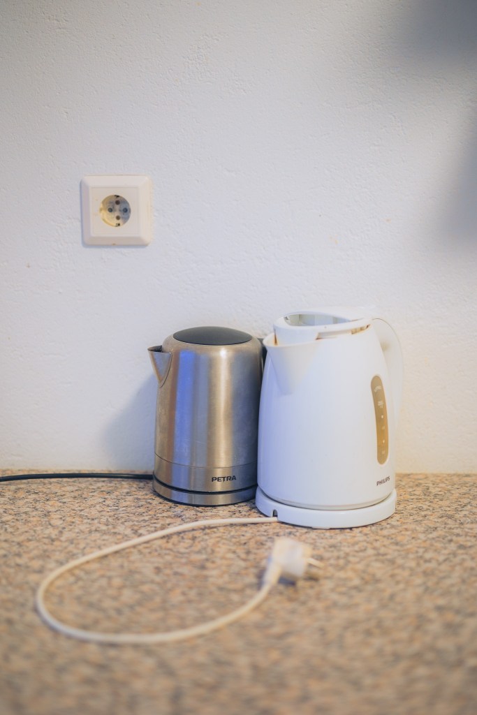Two electric kettles, one silver and one white, sit on a speckled countertop near a wall outlet. A power cord is loosely coiled in the foreground.