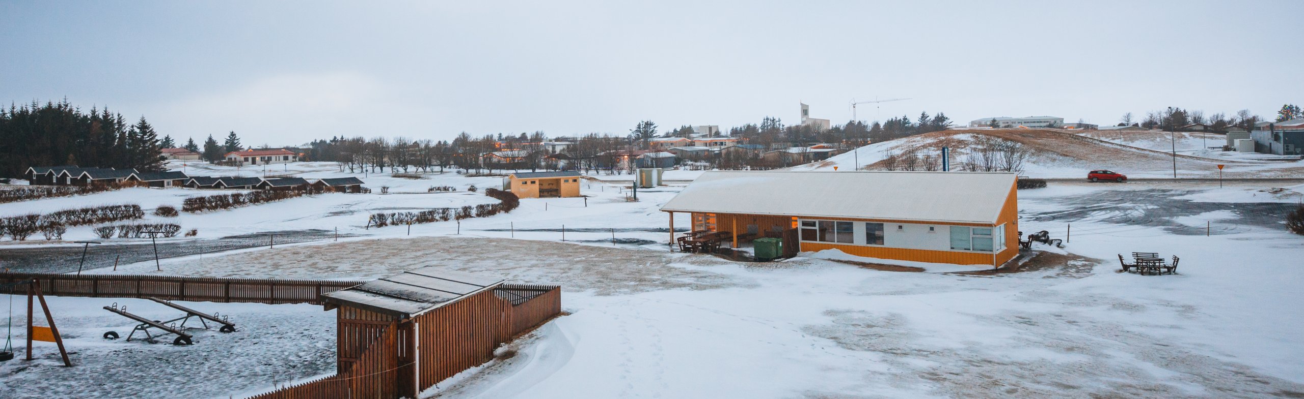 Snow-covered landscape with a yellow building and playground, including a seesaw and picnic tables.