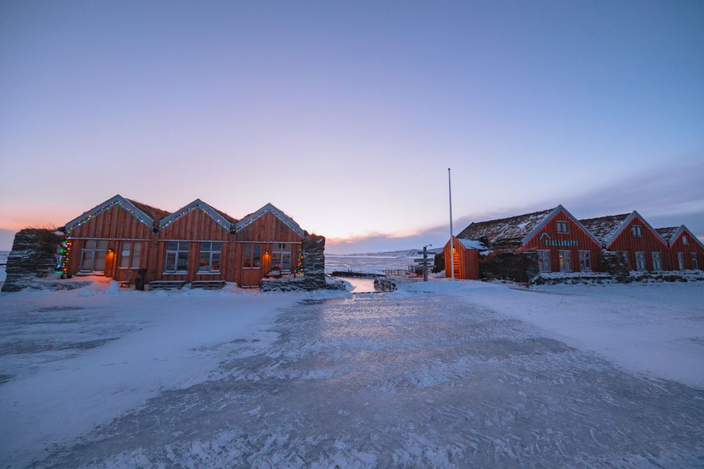 Snow-covered wooden cabins with festive lights at dawn. Soft purple and pink hues fill the sky.