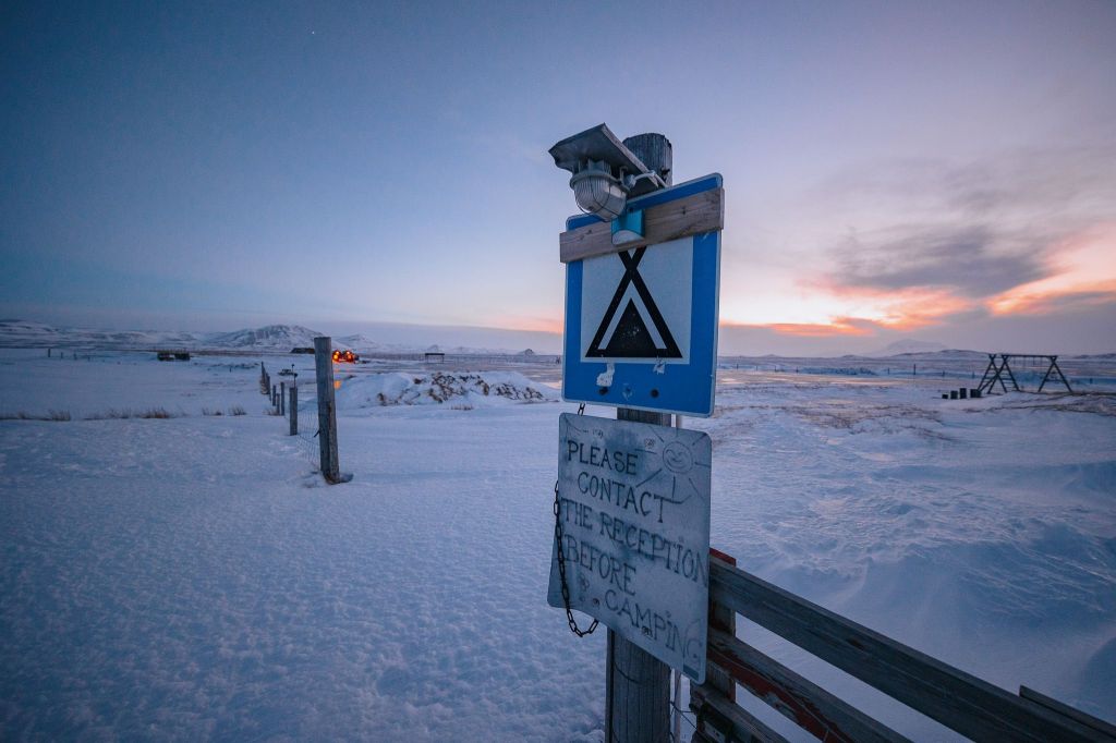 Snowy landscape at dusk with a blue camping sign and handwritten notice to contact reception before camping. A warm sunset glows in the background.