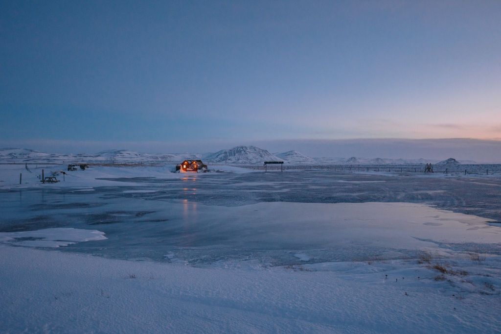A serene, icy landscape at dusk with a small, warmly lit cabin in the distance. Snow covers the ground, reflecting the soft, fading light of the sky.