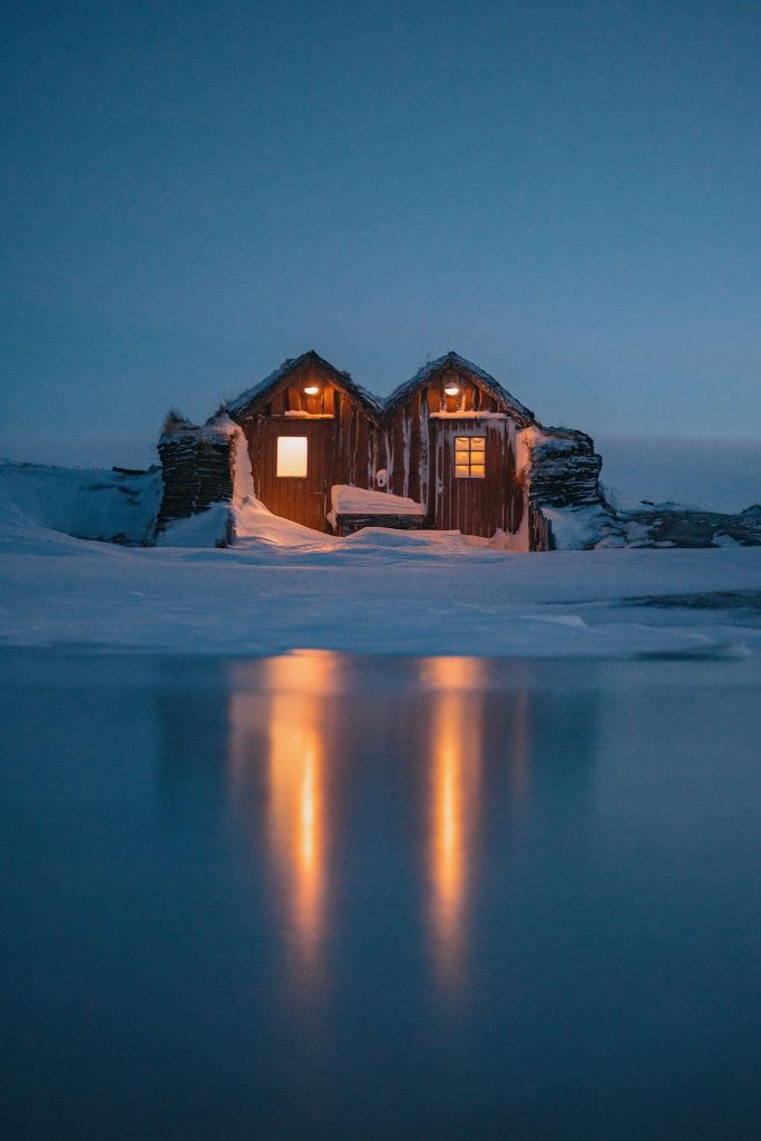 Two cozy cabins with warm lights glow in a snowy, twilight landscape. Their reflections shimmer on a smooth, icy surface.