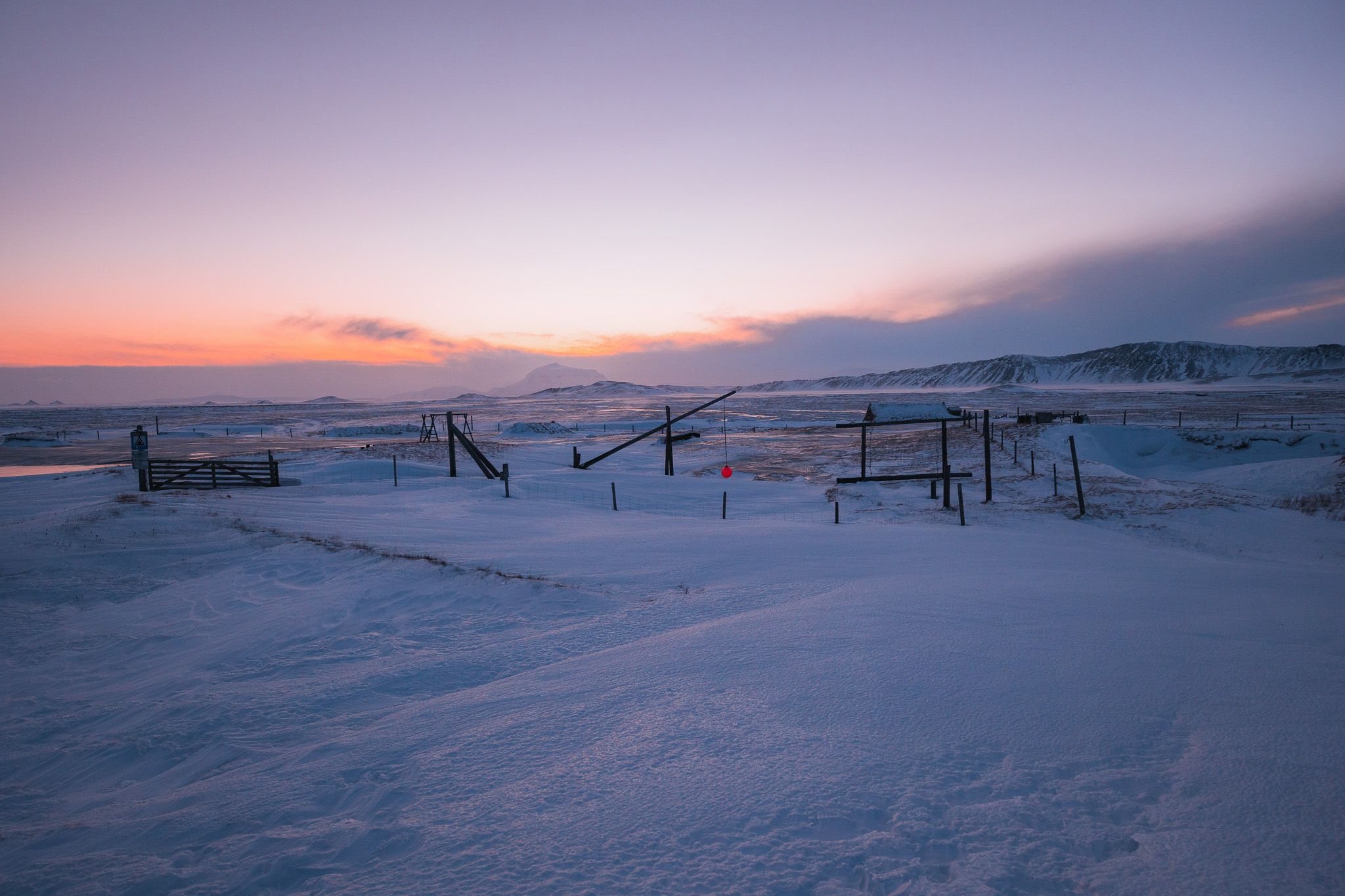 Snow-covered landscape at sunset with soft pink and orange hues in the sky. Silhouetted fences stand in the foreground.