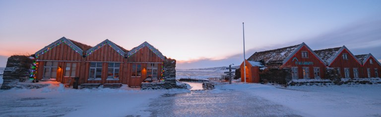 Cozy wooden cabins decorated with festive lights stand in a snowy landscape under a pastel sky.