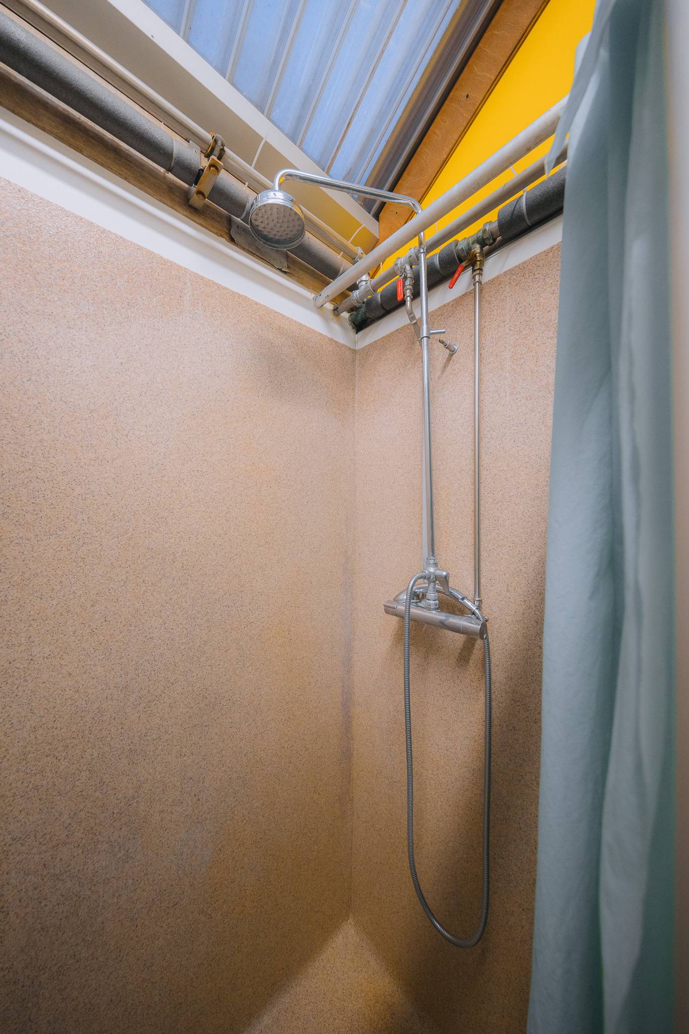 Small shower area with beige walls and a metal showerhead attached to overhead pipes. A pale blue curtain hangs partially open.