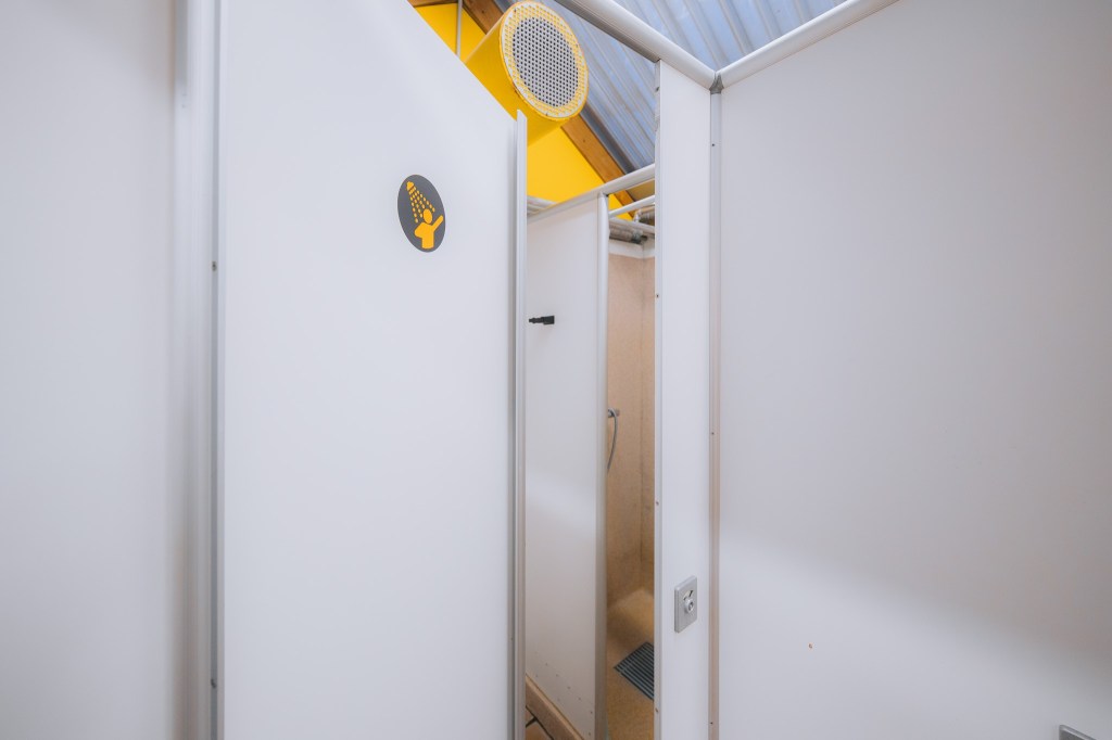 A clean shower stall in a bright space with white panels and a yellow accent wall. A round ventilation fan is visible above.