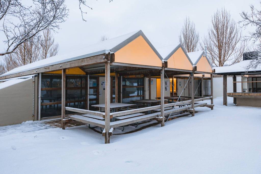 A snow-covered, open-air structure with three triangular roofs frames picnic tables.