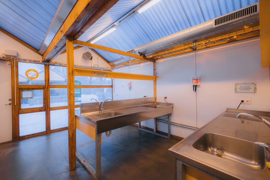 A clean, brightly lit communal kitchen with metal sinks, wooden beams, and a large window showing a snowy landscape outside.