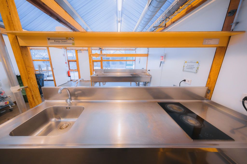 Stainless steel kitchen workspace with a sink and stovetop. Bright lighting, clean lines, and a "Keep clean!" sign above the sink.